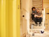 A young couple doing up their hallway in the old house they have bought . They are sitting on the dust sheets on the stairs and looking through the paint sample cards choosing the final colour for the beige hall .