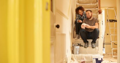 A young couple doing up their hallway in the old house they have bought . They are sitting on the dust sheets on the stairs and looking through the paint sample cards choosing the final colour for the beige hall .